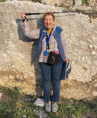 Norma Franklin demonstrating the height of the 4m - high artificial scarp separating the royal palace at Sebaste from the rest of the surrounding area . She is 1:50 meter s tall so ca 2 . 5 meters of the rocky scarp is now buried by earthen debris washed since the area was excavated in 1910. ( Photo by M . van den Berg )